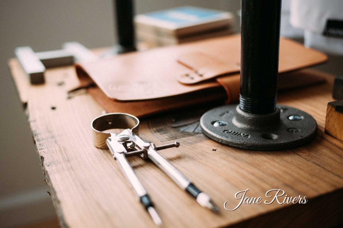 Drafting compasses and a leather folio on an artist's wooden desk, with a signature watermark in the corner