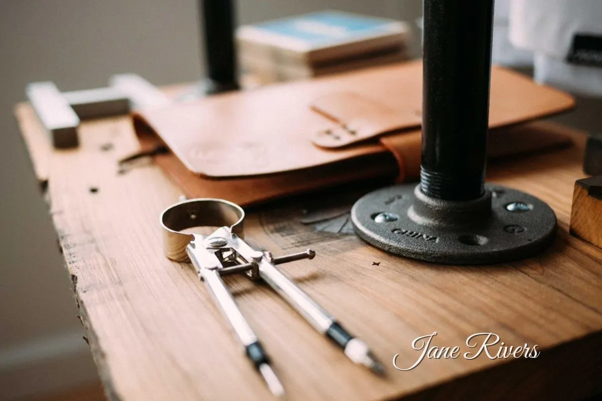 Drafting compasses and a leather folio on an artist's wooden desk, with a signature watermark in the corner