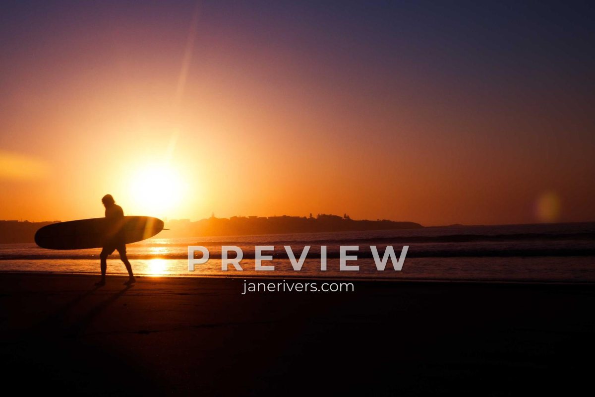 A surfer walking along the beach at sunset with PREVIEW and janerivers.com across the lower middle of the photo