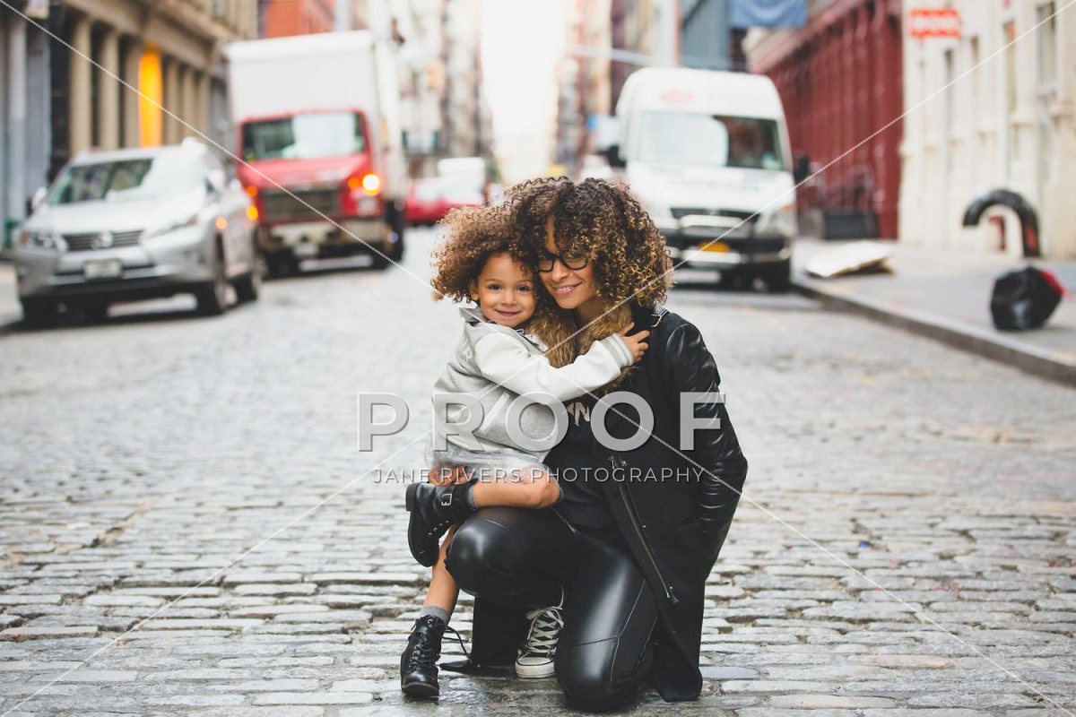 A mother hugging her child on a cobbled street, with a PROOF watermark over their legs and faint diagonal lines across the frame