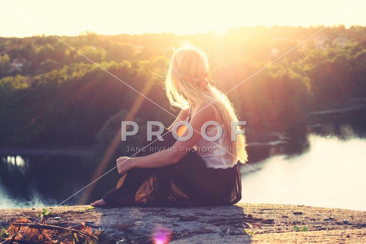 A woman sitting by a lake at sunset with a PROOF watermark and diagonal lines