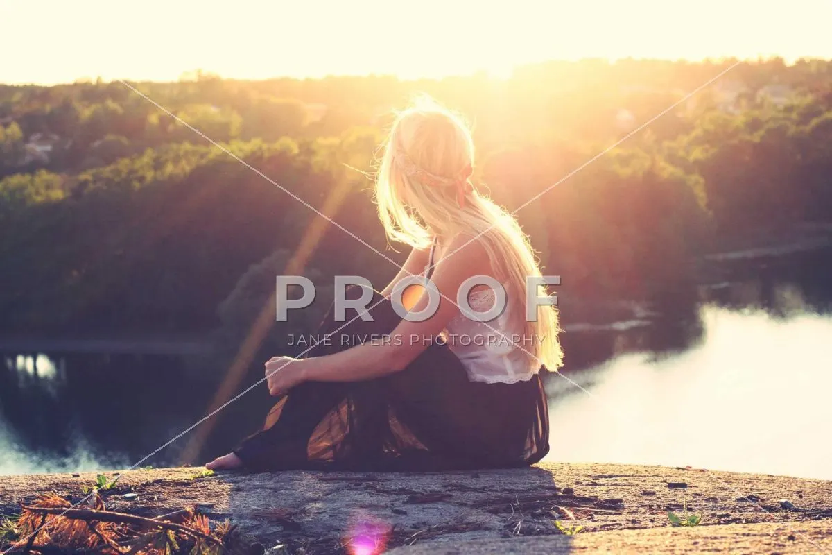 A woman sitting by a lake at sunset with a PROOF watermark and diagonal lines
