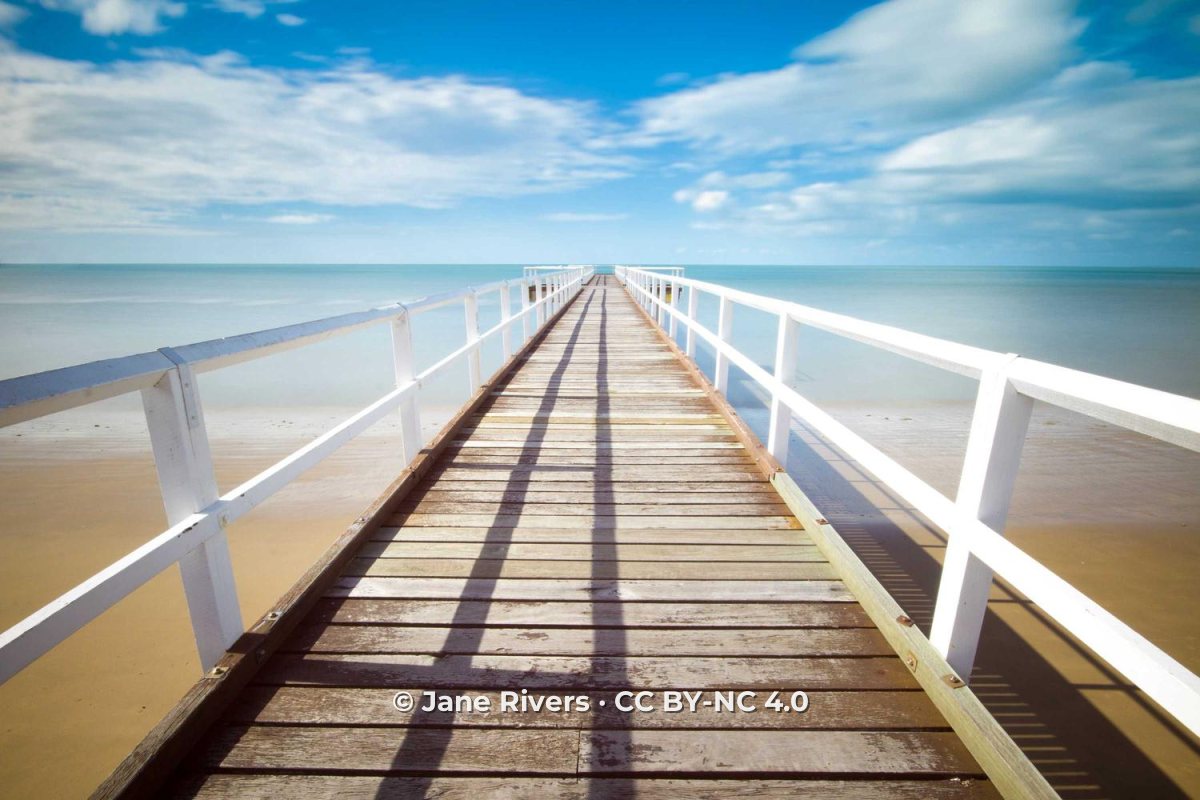 A long wooden pier stretching out over a turquoise sea, with a watermark reading © Jane Rivers · CC BY-NC 4.0