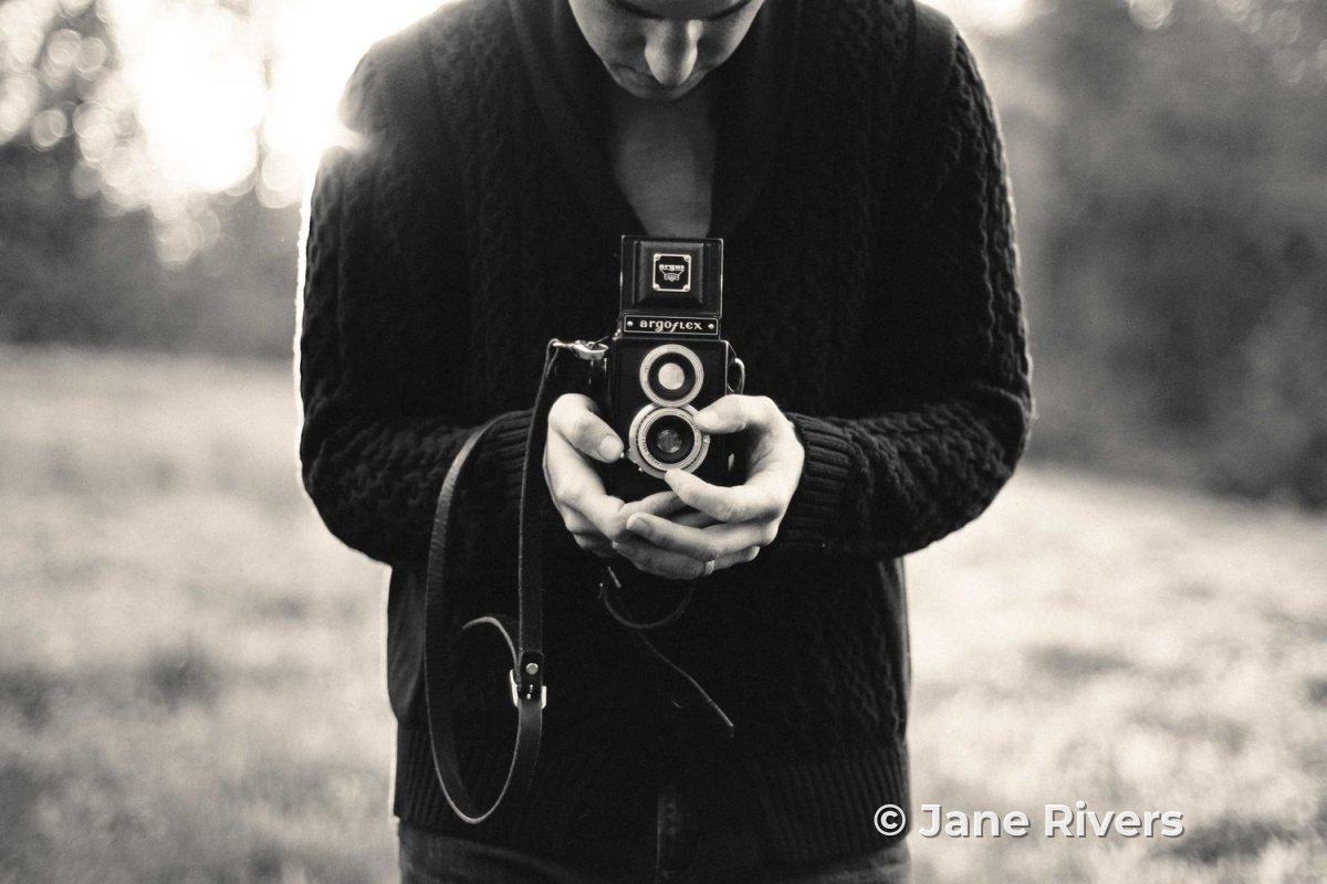 A black and white photo of a photographer holding a vintage twin-lens camera, with a copyright watermark