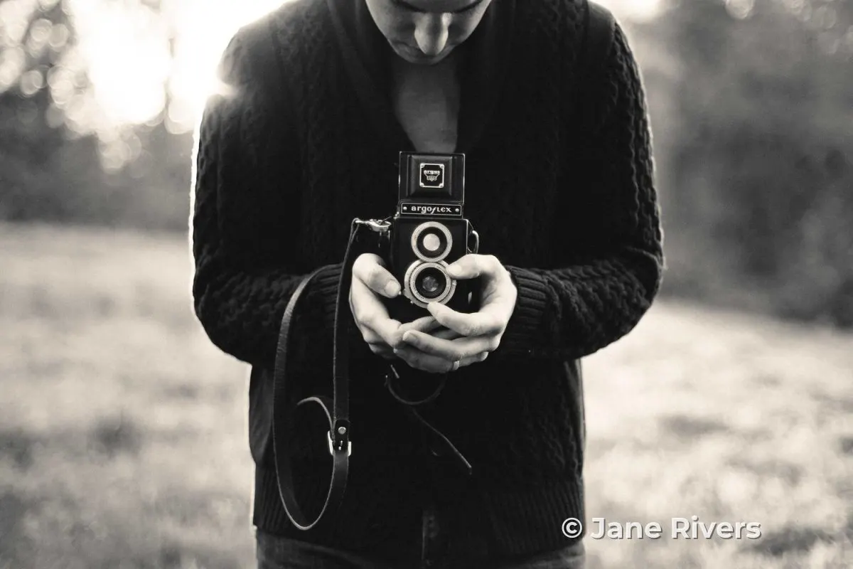 A black and white photo of a photographer holding a vintage twin-lens camera, with a copyright watermark