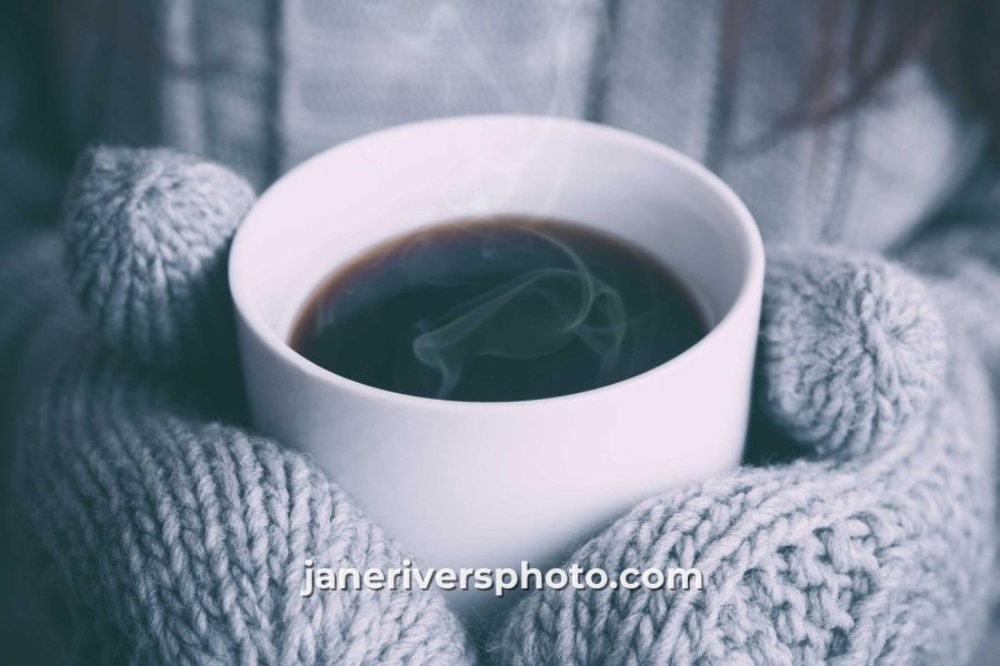 A cup of coffee held in knitted mittens with the watermark janeriversphoto.com