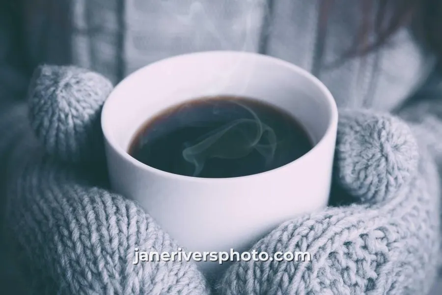 A cup of coffee held in knitted mittens with the watermark janeriversphoto.com