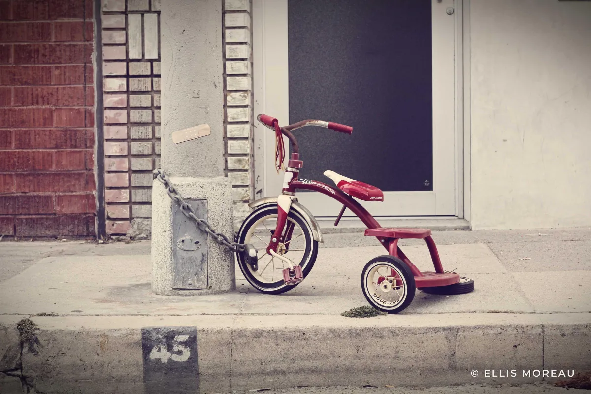 A red tricycle chained to a pole on a sidewalk, with a small © ELLIS MOREAU in white in the lower right corner over plain pavement