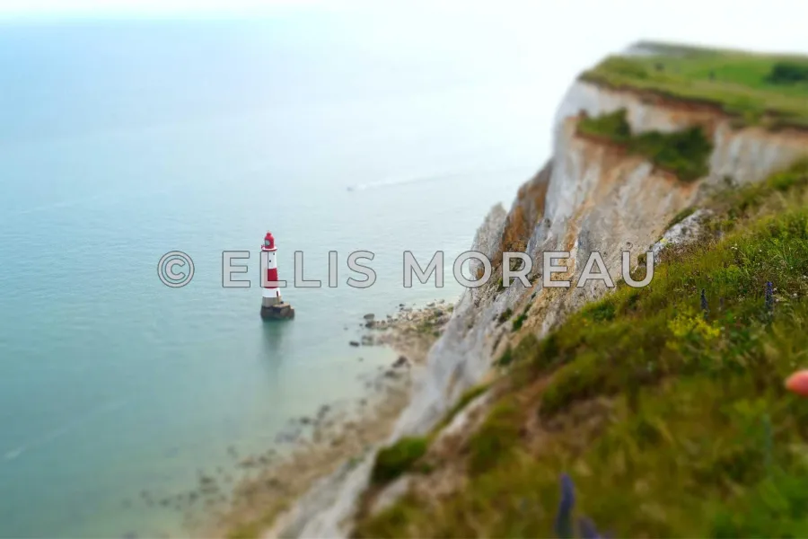 A lighthouse below a chalk cliff, with © ELLIS MOREAU in white letters with a dark outline, readable over both pale sea and dark grass