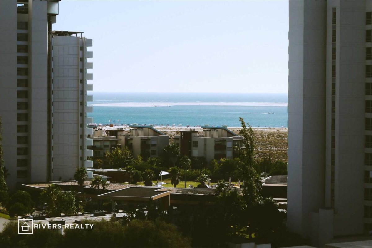 Apartment buildings framing a sea view, with a small agency logo reading RIVERS REALTY in the lower left