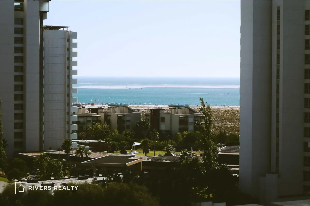 Apartment buildings framing a sea view, with a small agency logo reading RIVERS REALTY in the lower left