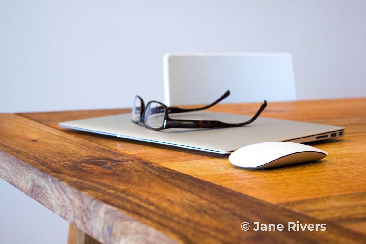 A laptop with glasses and a mouse on a wooden desk, with a small copyright watermark