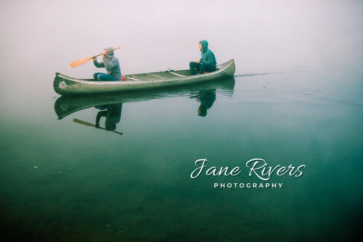 Two people in a canoe on misty green water, with a signature and the word Photography placed below and to the right of the canoe