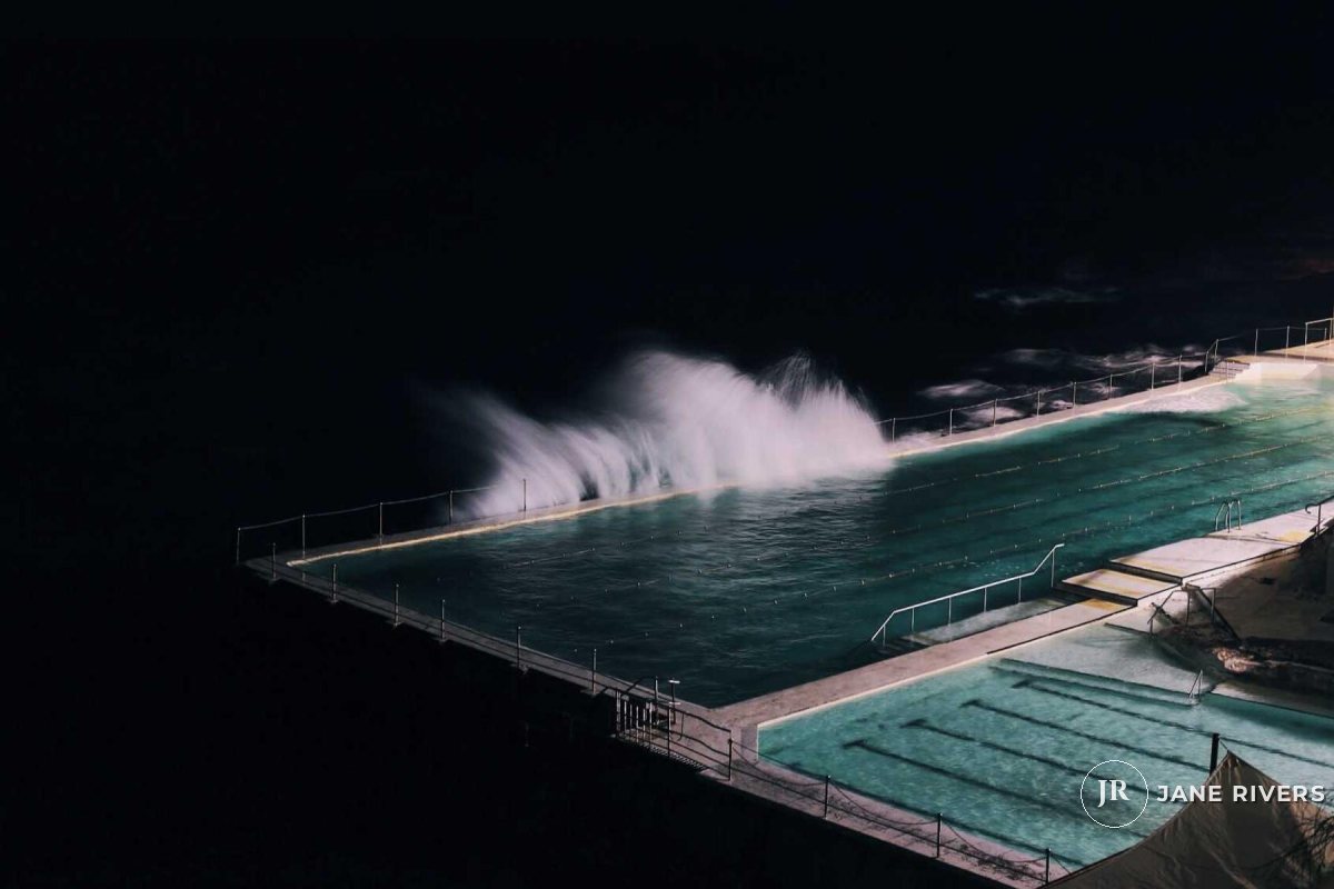 A video frame of a wave breaking over a sea pool with a small monogram and name in the bottom-right corner