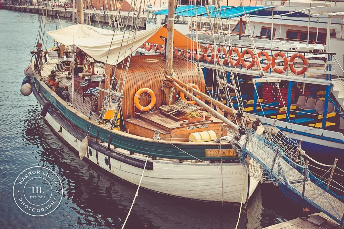 A wooden sailing boat in a harbor with a round white badge over the water in the lower left: HARBOR LIGHT curving along the top, PHOTOGRAPHY along the bottom, and HL with EST. 2019 in the middle