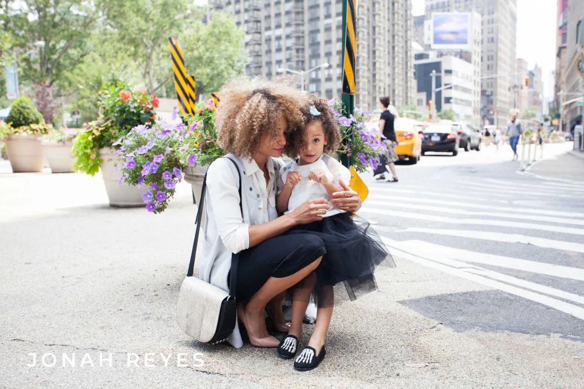 A mother and daughter crouching on a bright city sidewalk with JONAH REYES in white letters that almost disappear against the pale pavement