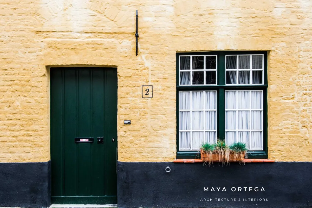 A yellow brick house with a dark green door and window, with MAYA ORTEGA, a short thin line and ARCHITECTURE & INTERIORS stacked in white on the dark base of the wall