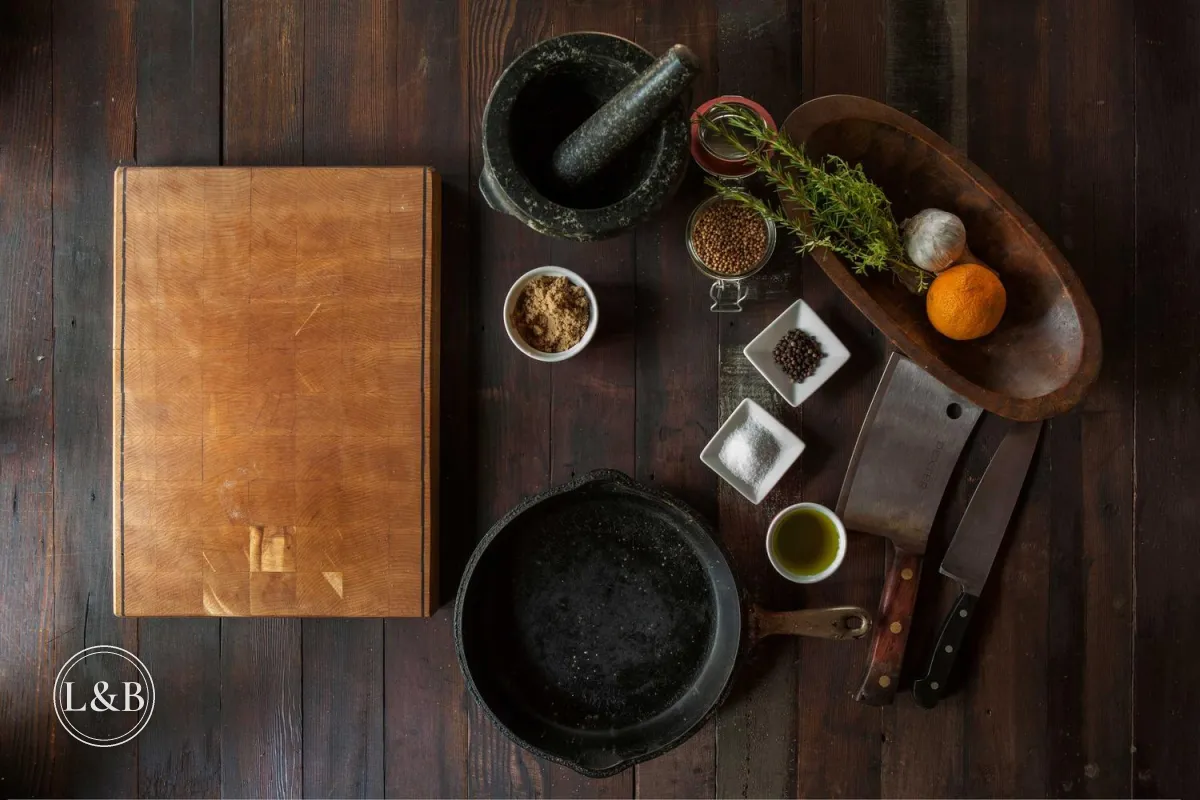 A rustic kitchen flat lay with a cutting board, cast iron pan, spices and knives on dark wood, with an L&B monogram in a thin double circle in the lower left