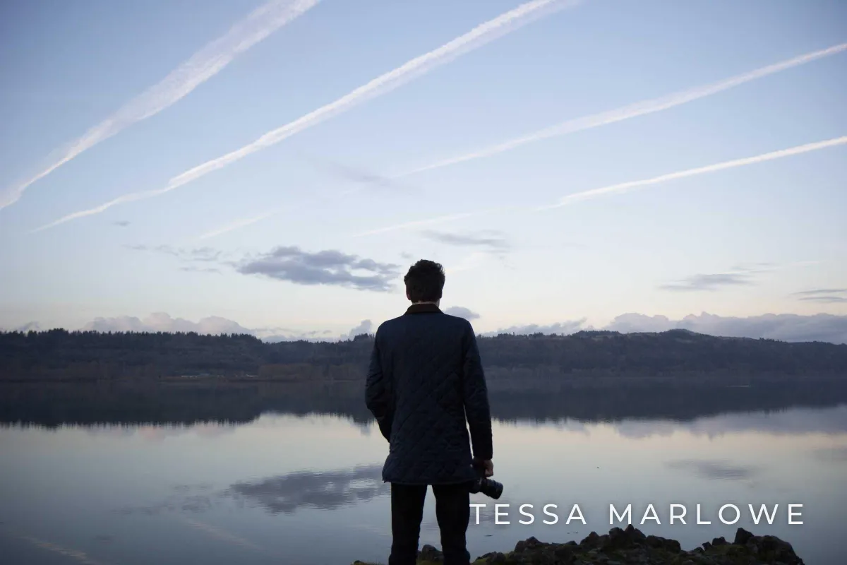 A man with a camera standing at a calm lake at dusk, with TESSA MARLOWE in small, widely spaced white capitals in the lower right