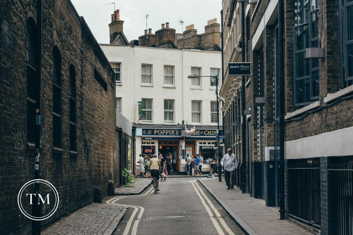 A narrow London street with brick walls and a small white TM monogram inside a double ring in the lower left