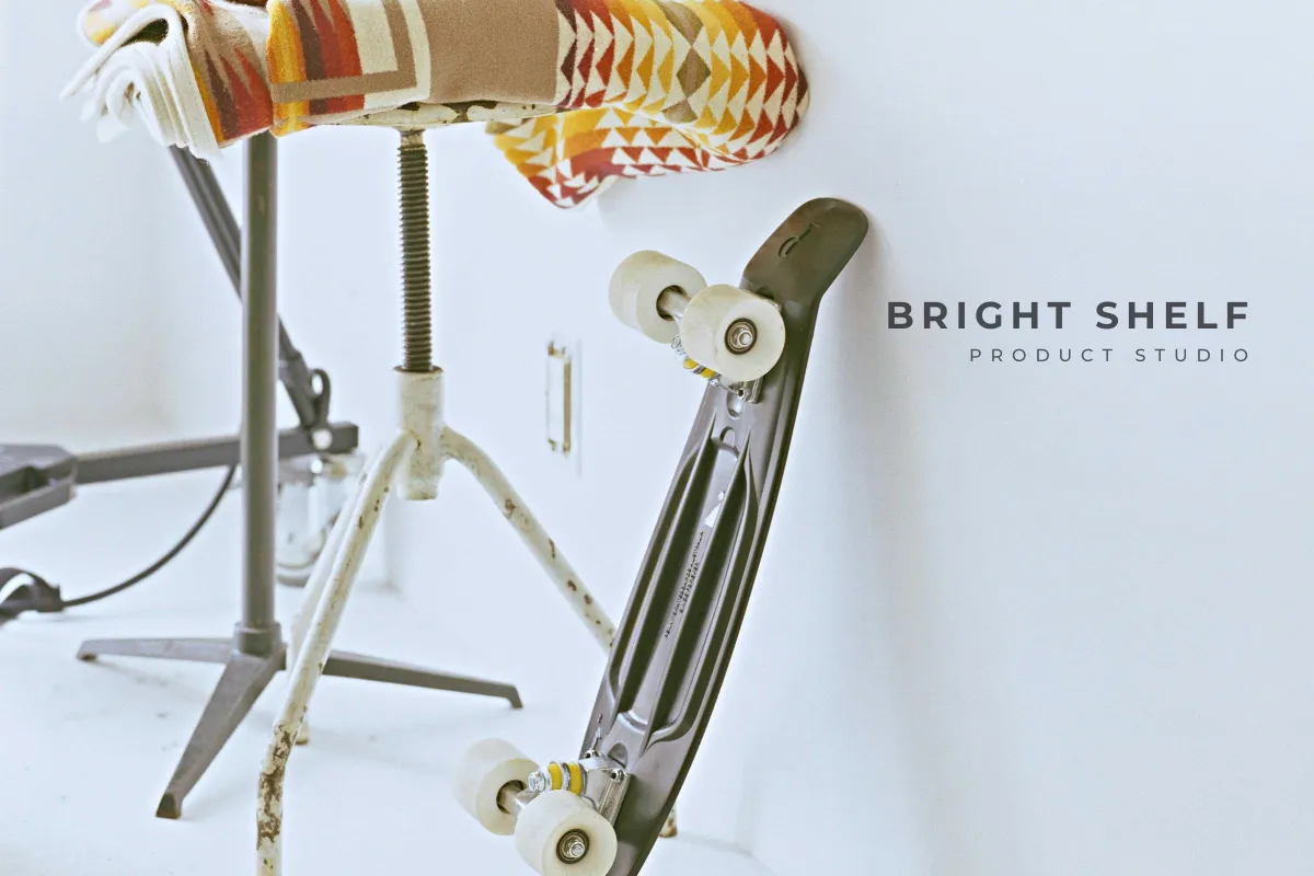A skateboard leaning on a white wall beside a stool, with BRIGHT SHELF and PRODUCT STUDIO in dark capitals on the wall