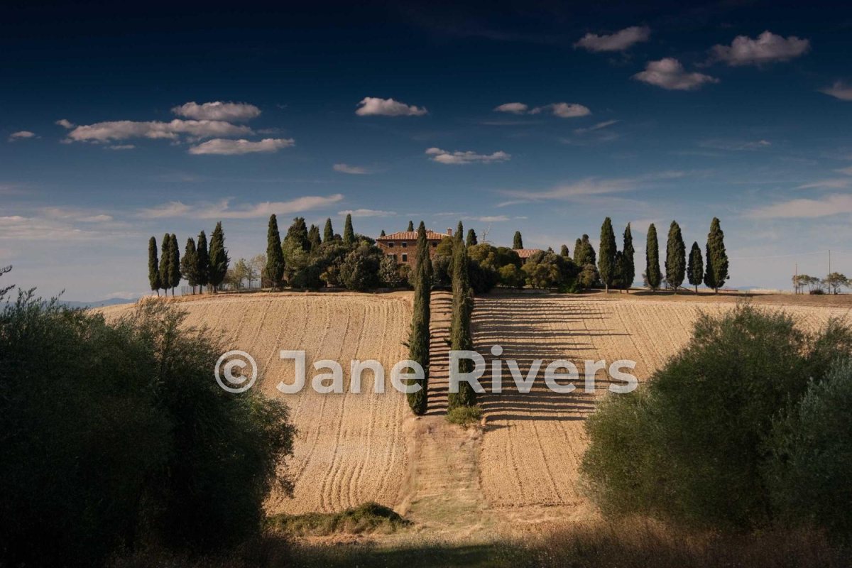 A Tuscan farmhouse on a hill with a large semi-transparent Jane Rivers watermark across the field
