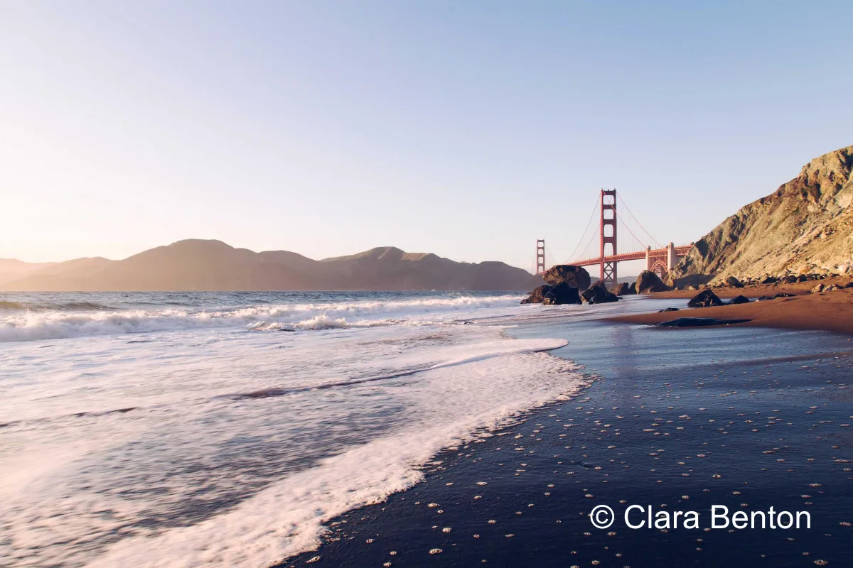 A beach at golden hour with a suspension bridge in the distance and © Clara Benton in plain solid white text in the lower right corner