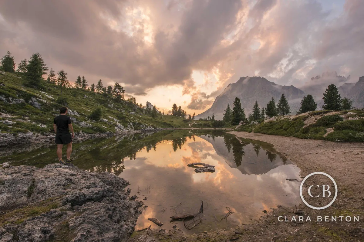 A hiker beside a mountain lake reflecting a pink sunset sky, with a white CB monogram ring and CLARA BENTON at about 70% opacity in the lower right corner