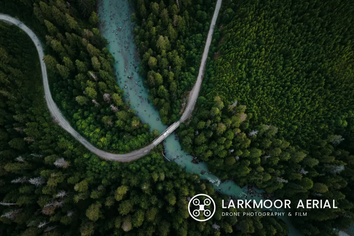 A top-down drone photo of a gravel road curving over a turquoise river through dense forest, with a white logo of a drone in a ring and LARKMOOR AERIAL, DRONE PHOTOGRAPHY & FILM in the lower right