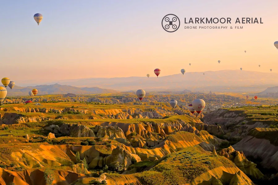 Hot air balloons over eroded valleys at sunrise with a charcoal drone logo and LARKMOOR AERIAL in the pale sky at the upper right