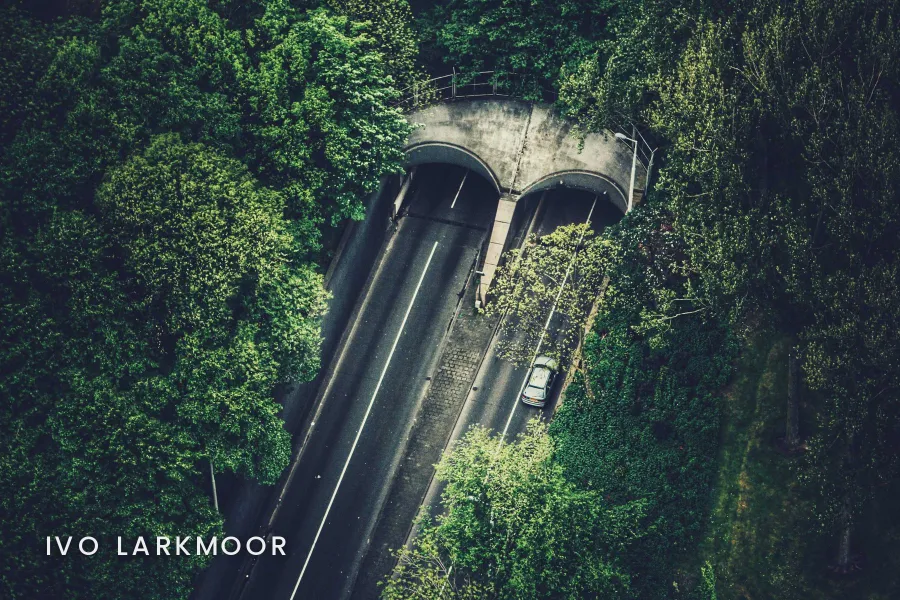 A drone view of a two-lane road entering twin tunnels under a forested hill, with IVO LARKMOOR in small spaced white capitals in the lower left