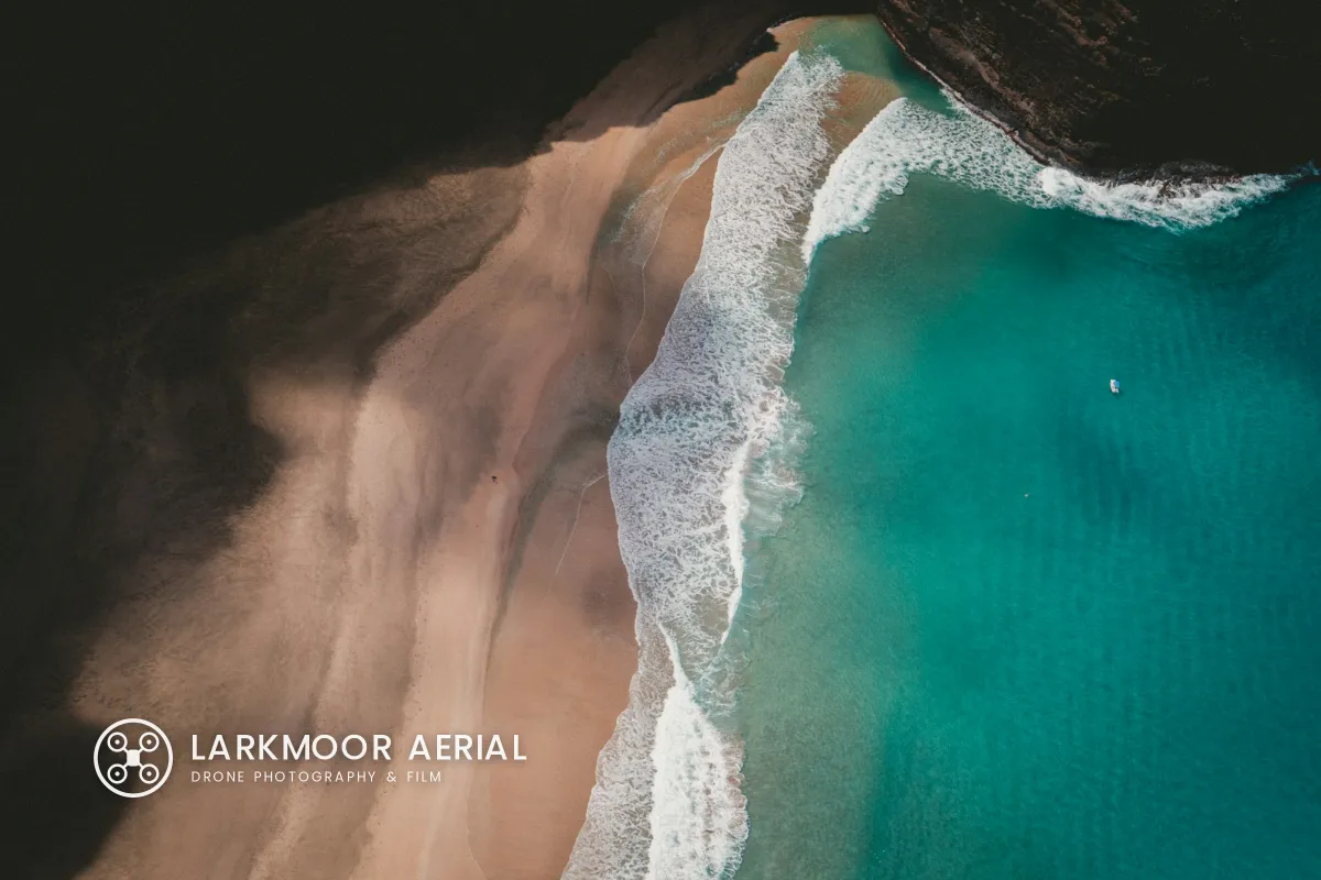 A top-down drone photo of a beach where sand meets turquoise sea and white surf, with a white drone logo and LARKMOOR AERIAL on the smooth sand in the lower left