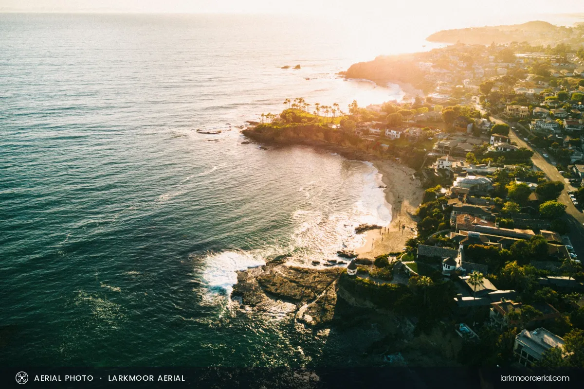 An aerial photo of a rocky cove and a hillside town at golden hour, with a slim dark band along the bottom reading AERIAL PHOTO, LARKMOOR AERIAL on the left and larkmooraerial.com on the right