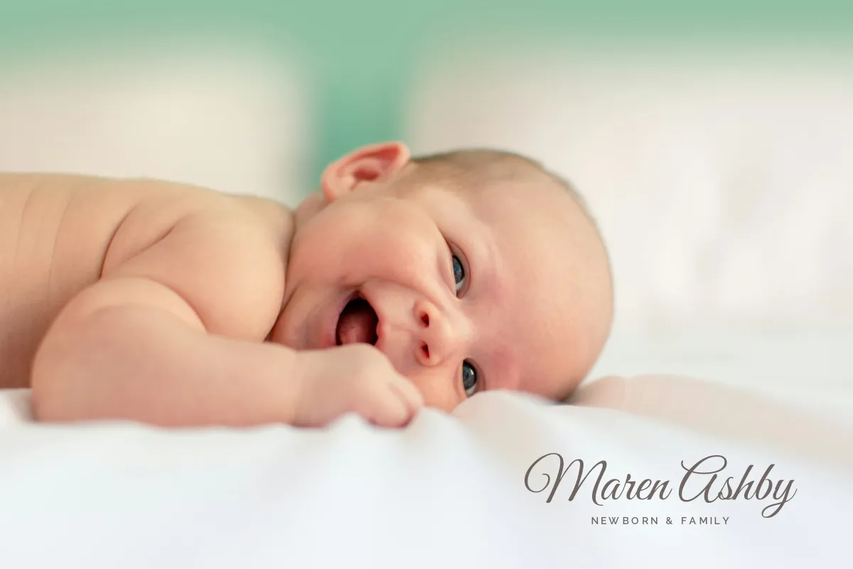 A smiling newborn lying on a white sheet against a soft mint background, with Maren Ashby in a warm gray script signature and NEWBORN & FAMILY in small capitals on the sheet in the lower right