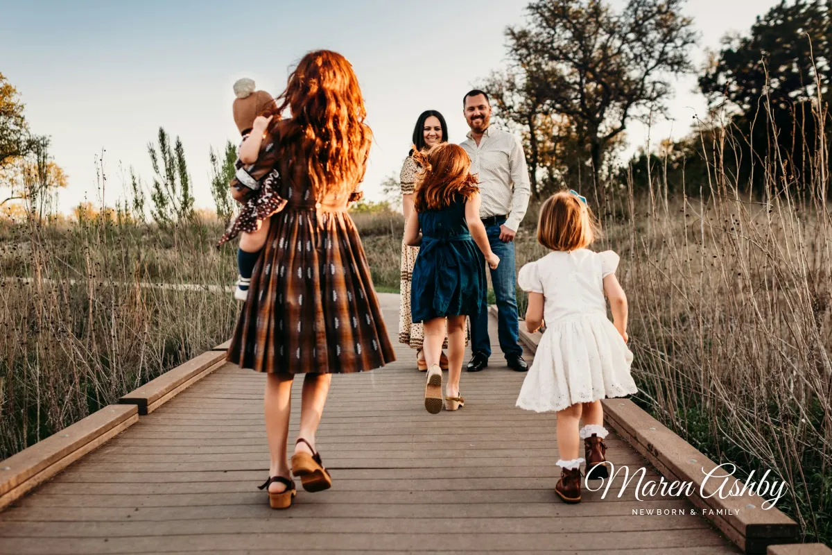 A family on a wooden boardwalk at golden hour, the mother carrying a baby and two girls running toward their parents, with a white Maren Ashby signature in the lower right