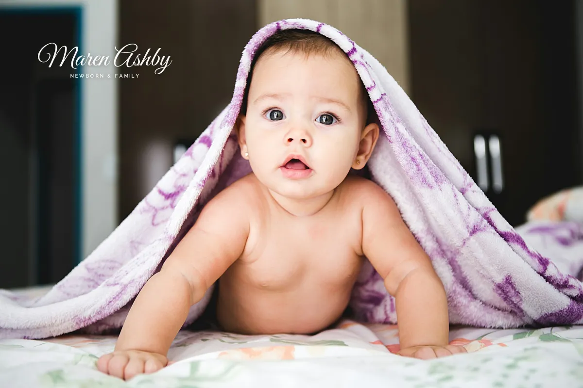 A baby peeking out from under a lilac blanket on a bed, with a small white Maren Ashby signature in the dark area at the upper left